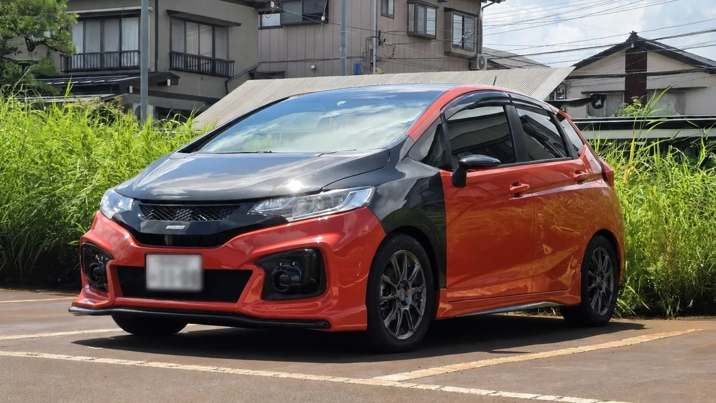 Un Honda Fit RS japonés anaranjado con bonete y guardalodos delanteros de fibra de carbono, estacionado en Japón con la tablilla borrosa.