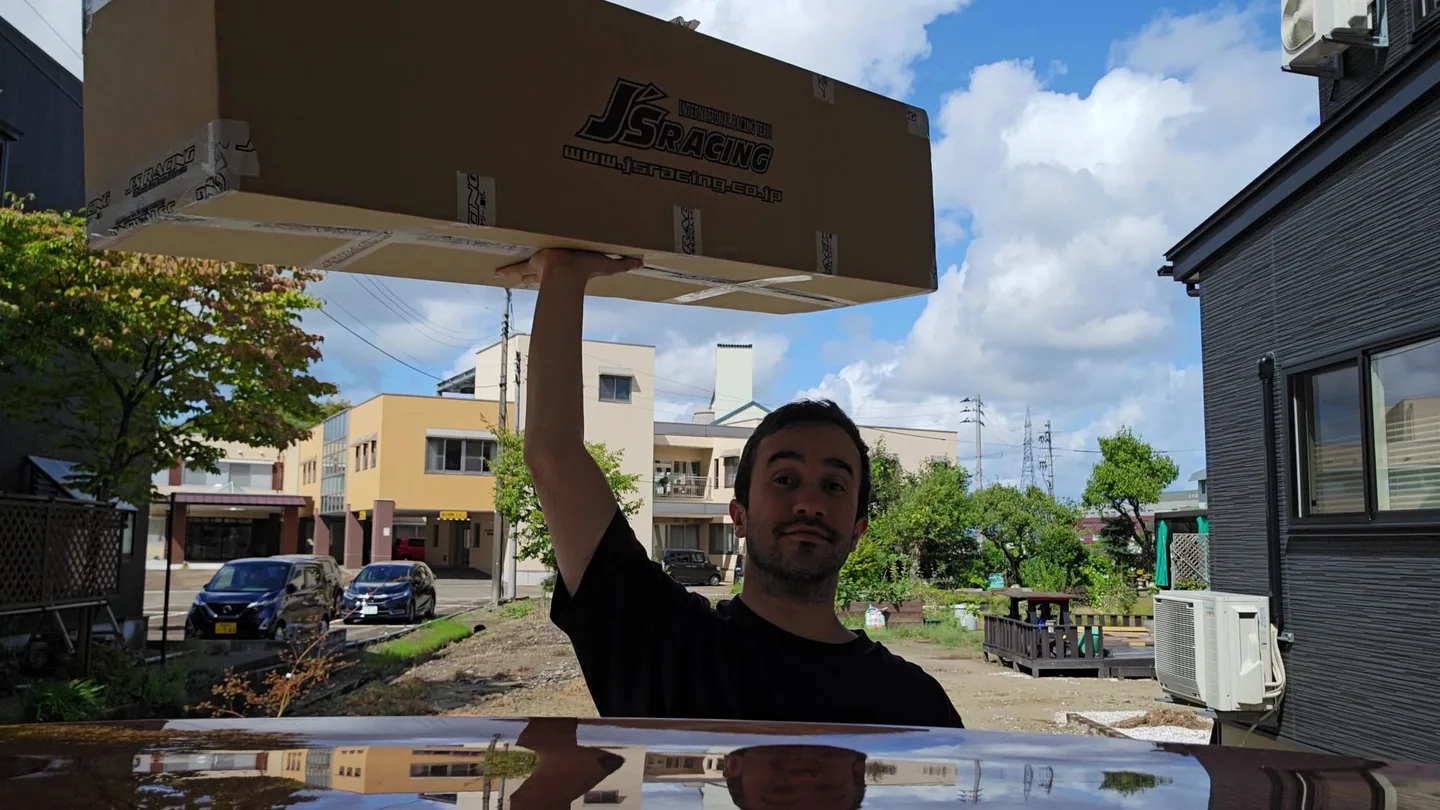 Harry sostiene una caja larga de J's Racing sobre su cabeza al lado del Honda Fit.