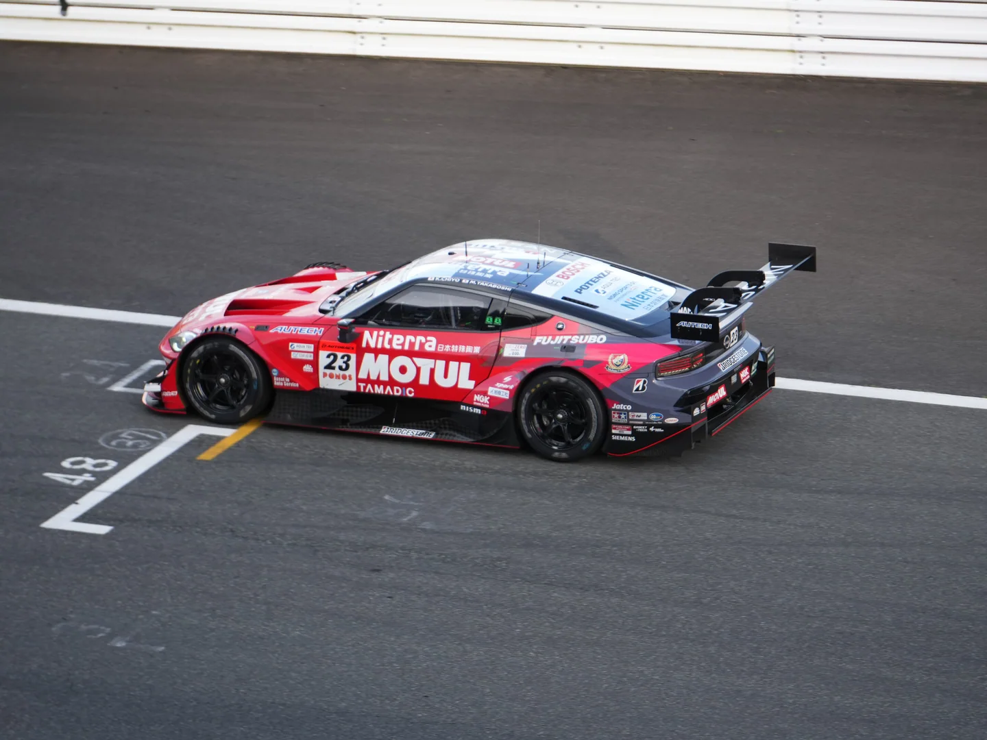 The red and black No. 23 MOTUL Niterra Nissan Z passes beneath the grandstand at Fuji Speedway.