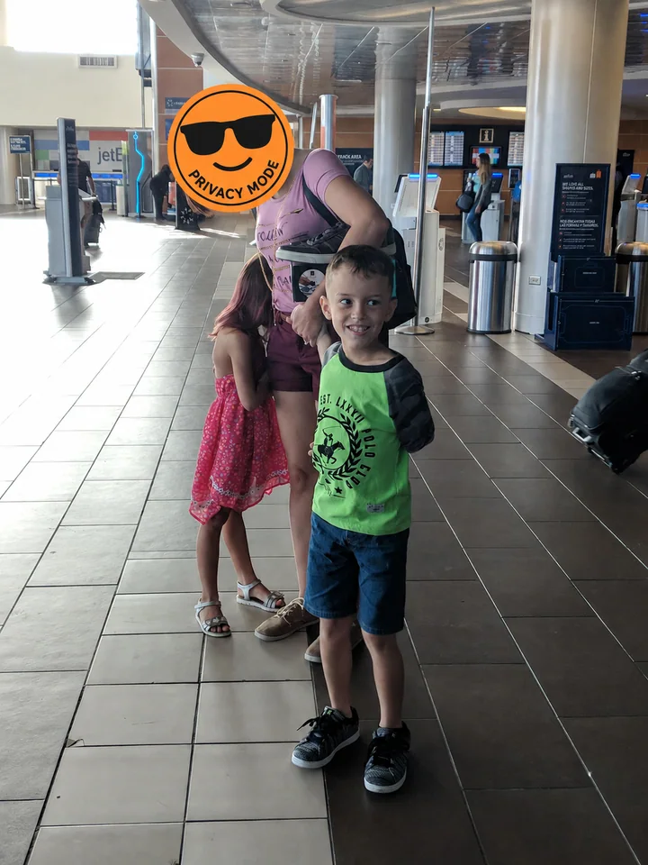 Two children stand with their mother at an airport farewell; an orange Privacy Mode stamp completely covers the adult woman's face.