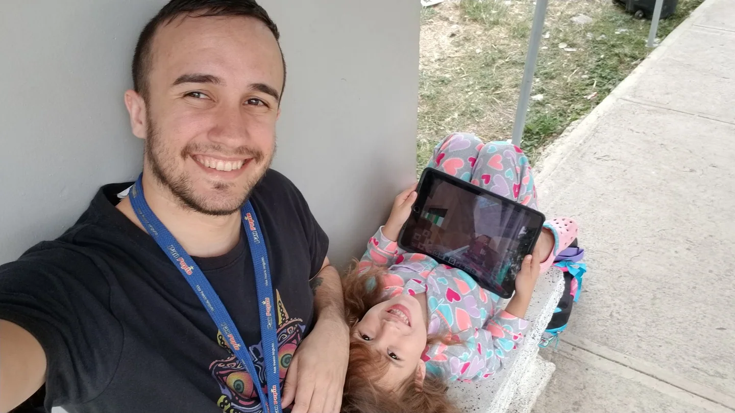 Harry smiles beside his daughter as she plays Minecraft on a tablet while lying upside down on a bench.