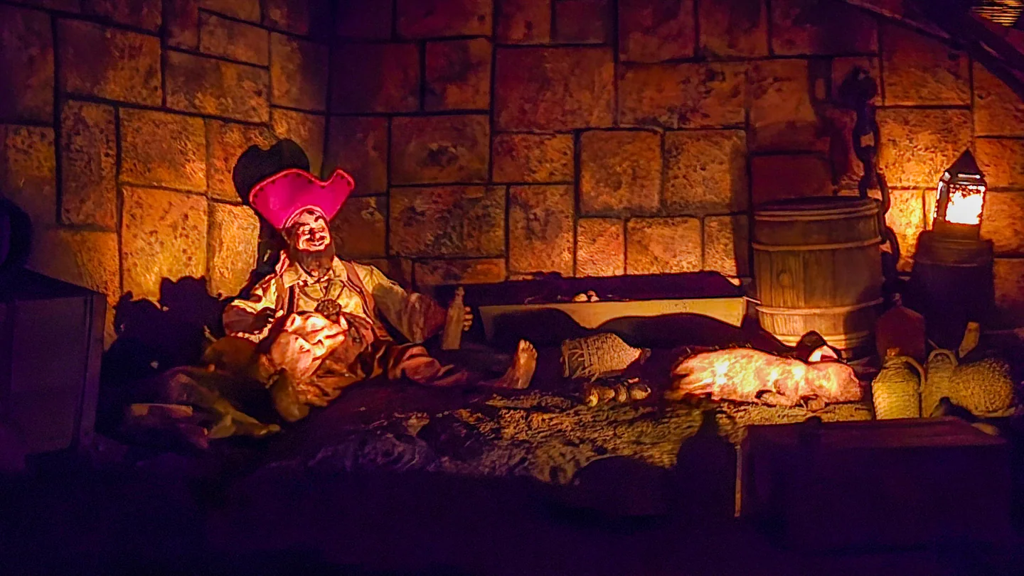 Un pirata animatrónico descansa junto a barriles y sacos bajo una luz cálida dentro de Pirates of the Caribbean.